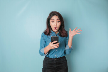 Surprised Asian woman wearing blue shirt pointing at her smartphone, isolated by blue background