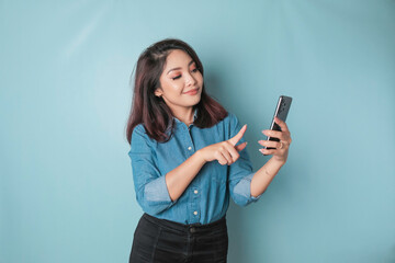 A portrait of a happy Asian woman is smiling and holding her smartphone wearing a blue shirt isolated by a blue background