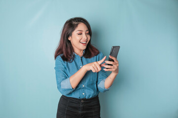 Fototapeta premium A portrait of a happy Asian woman is smiling and holding her smartphone wearing a blue shirt isolated by a blue background