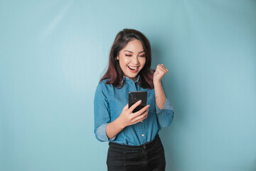 A young Asian woman with a happy successful expression wearing a blue shirt holding smartphone...