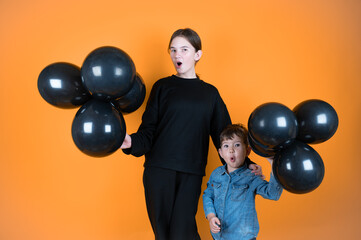 Surprised teengirl and little boy holding black balloons in hands. Orange bright background. Sales concept