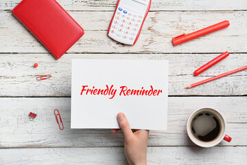 Human hand holding paper sheet with text FRIENDLY REMINDER on white wooden background