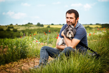 Naklejka na ściany i meble Tranquil and cheerful scene with a white Caucasian bearded man sitting outside in the nature, looking dreamy and hugging his best friend dog.