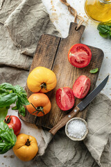 Raw tomatoes and knife on wooden cutting board surrounded by ingredients salt basil leaves and olive oil around. Top view. Rustic cooking background
