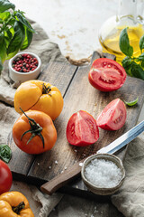 Raw tomatoes and knife on wooden cutting board surrounded by ingredients salt basil leaves and olive oil around. Rustic cooking background