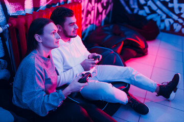 Young man and woman couple plays on game console with gamepad sitting on chair.