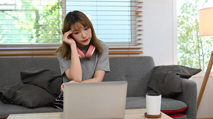 Serious young woman reading email, working online from home with laptop on couch 