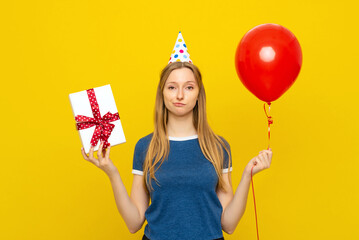 Sad young woman holds box with gift present and red inflated helium balloon, has bad mood during celebration, wears party cone and blue t shirt, feels embarrassed