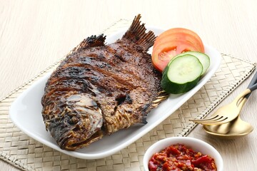 Gurame Bakar Madu, Grilled Gourami with honey and soy sauce. Served with sambal (chilli sauce), cucumber and tomatoes in white plate.
