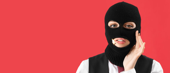 Portrait of young woman in balaclava and with burning match in mouth on red background with space...