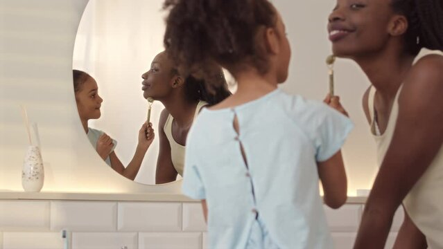 Medium slowmo of young beautiful Black woman and her 6 year old daughter using quartz roller while doing face massage to each other standing in front or mirror in bathroom