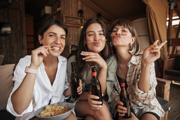 Positive young caucasian girls grimacing at camera, relaxing with drink in country house. Brunettes wear casual clothes. Mood, lifestyle, concept.
