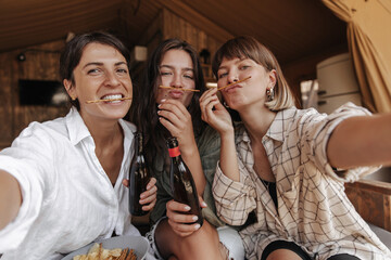 Positive young caucasian girls grimacing looking at camera spending leisure time together. Brunettes are drinking and celebrating weekend. Concept of enjoying moment