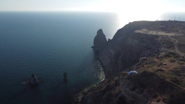Aerial drone view of a man flying a white and blue paraglider over a hill and trees to the sea waves near the rocks. Active paraglider flight over the seascape with clear skies at suset. Extreme sport