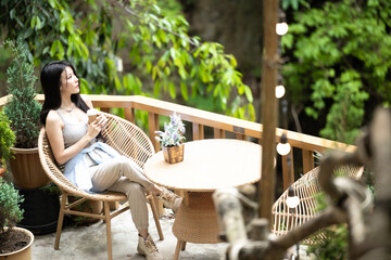 Beautiful young asian woman holding a cup of coffee sitting on a chair and look out to nature.  Young asian woman drinking coffee and relaxing in the midst of nature. Beautiful nature. - Powered by Adobe