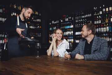 Happy European heterosexual man and young woman date restaurant, sommelier pours red wine from...
