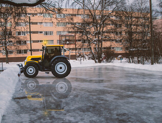 A yellow tractor removing snow in the playground ice of a vintage living district in winter with a retro concrete block house in the background and copy space