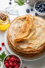 Stack of French crepes or traditional Russian blini on white plate on light background. Thin pancakes served with raspberries, blueberries and honey. Summer concept. Selective focus.