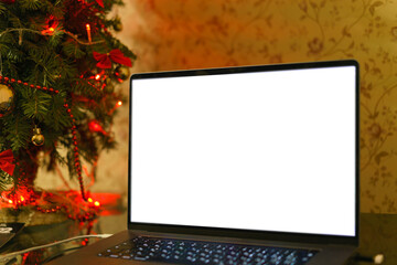 A laptop with an empty mockup display stands on a table with small Christmas on the background. Keyboard. Ribbon. Work. Person. Decor. House. Apartment. Browsing. Cozy. Consumer. Client. Working. Fest