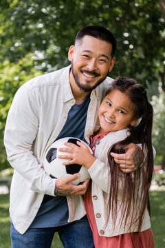 Positive asian dad and girl holding soccer ball and looking at camera in park.