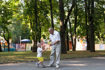 Fototapeta premium Grandfather and grandson are walking in the park along the path with a glass of popcorn. An old man and a little boy have snacks on the go.