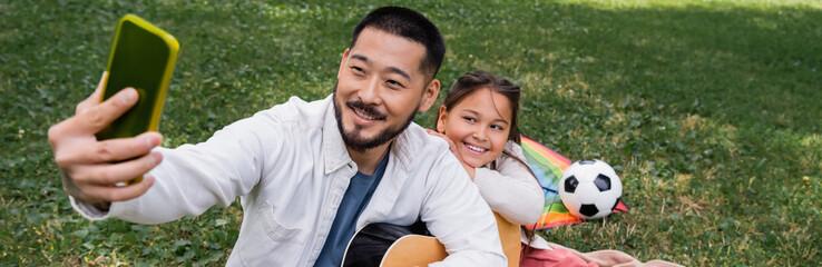 Fototapeta premium Asian dad taking selfie on cellphone while holding acoustic guitar near child in park, banner.