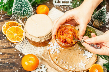 Woman's hands hold orange marmalade or orange jam in a glass jar. Sweet confiture with festive Christmas decor.