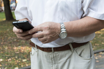 Fototapeta na wymiar A mobile phone in the large hands of an old man with a watch on his left hand, in a white shirt, on a walk in the park