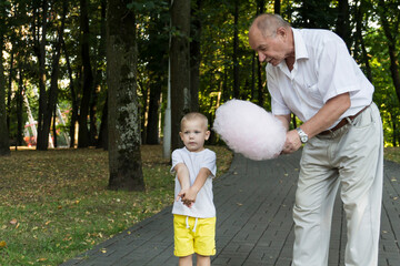 Grandfather lovingly and carefully treats and feeds his little grandson with pink cotton candy in an amusement park. A pensioner and a little boy spend their free time on weekends.