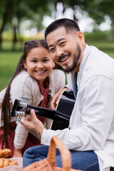 Positive asian man playing acoustic guitar and looking at camera near daughter in park.