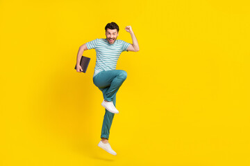 Fototapeta na wymiar Full length photo of excited handsome man dressed blue pants win sports betting hold laptop fist up isolated on yellow color background