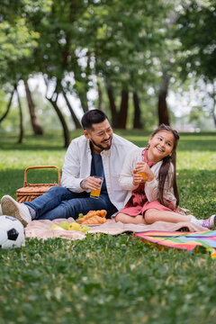 Happy asian family holding orange juice near food and football in summer park.