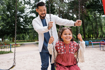Smiling father standing near asian daughter on swing in park.