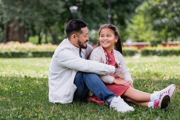 Asian parent hugging happy preteen daughter on grass in park.