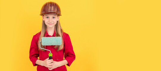 smiling teen child in builder hard hat with paint roller on yellow wall. Child builder in helmet horizontal poster design. Banner header, copy space.