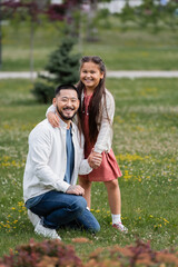 Fototapeta premium Cheerful asian kid hugging father on meadow with flowers in park.