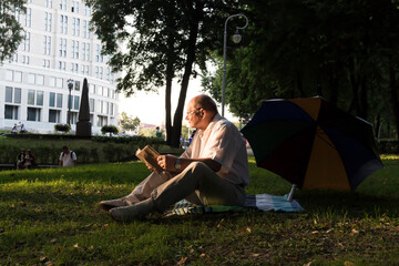 An elderly man in a white shirt is sitting on a blanket, on the ground in a park and reading an...