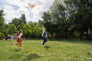 Asian father holding flying kite while running with daughter in park.