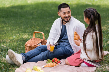 Smiling asian parent holding orange juice near daughter with croissant in park.