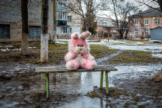 Pink plush rabbit sits on a bench in a poor residential area. Bad wheather. Deppresion. Sadness