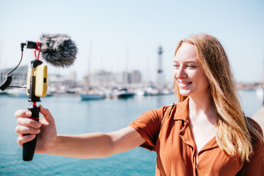 female travel vlogger streaming near sea in a touristic destination. She looks to her smart phone smiling