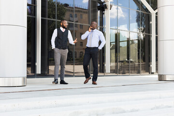 Portrait of two dark-skinned businessmen walking and talking in front of a modern building exterior. Friendly meeting outdoors