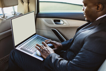 Business concept of a handsome dark-skinned man in a suit and glasses in the back seat of a luxury car working with a laptop. Copyspace and mockup