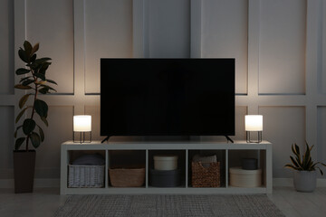 Modern TV on cabinet, lamps and beautiful houseplants near white wall in room. Interior design