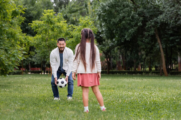 Fototapeta premium Smiling asian dad holding football near daughter in summer park.