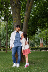 Naklejka na ściany i meble Smiling asian dad hugging and looking at daughter near tree in park.