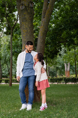 Smiling asian dad hugging and looking at daughter near tree in park.