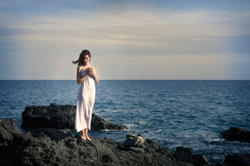 Fototapeta na wymiar Feminine woman standing on rocky seashore at sundown