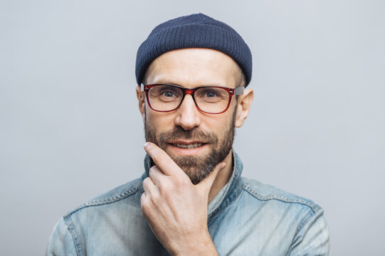 Confident satisfied bearded male keeps hand on chin, listens attentively interlocutor, wears fashionable clothing, isolated over white studio background. People and facial expressions concept