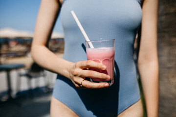 Closeup of a woman wearing blue swimsuit, holding a glass with strawberry milkshake at the beach.